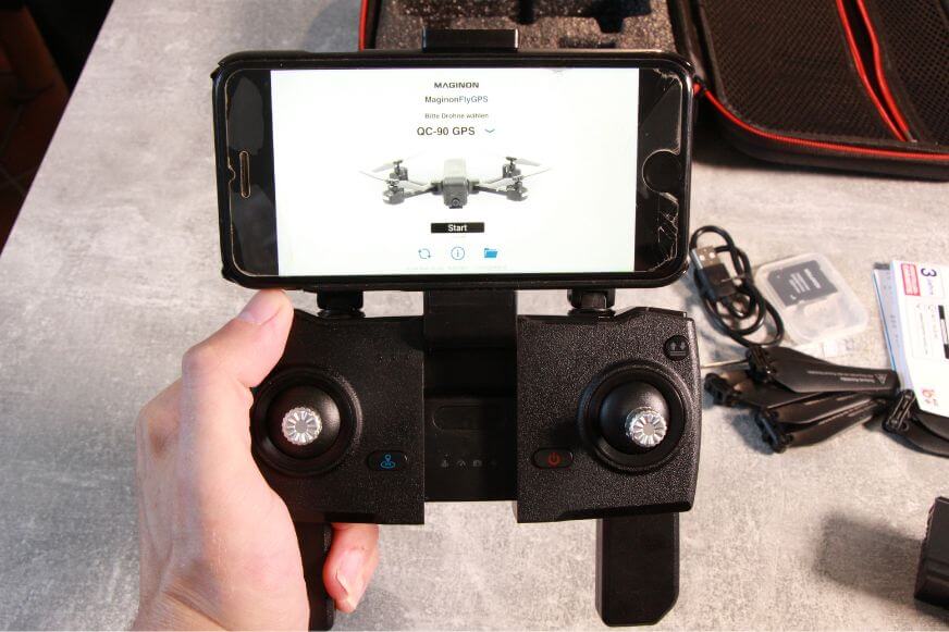 Das Smartphone ist im Controller der Maginon QC 90 Drohne eingelegt und die Drohne liegt dahinter auf dem Tisch.